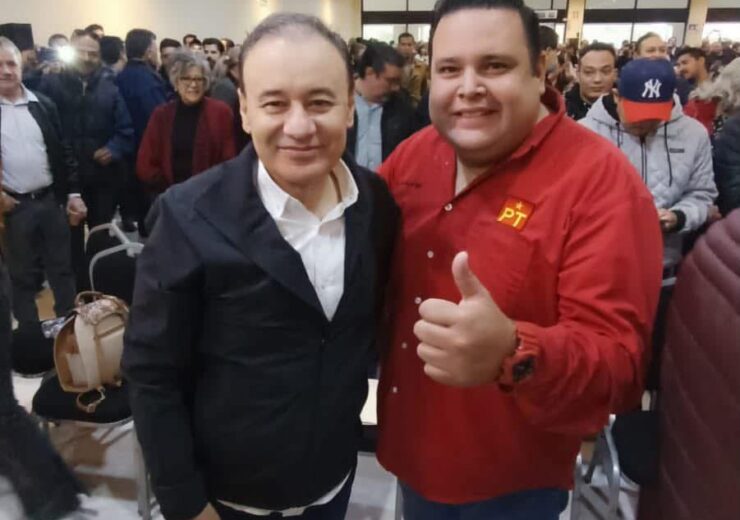Dos asistentes posan durante un encuentro político en Hermosillo; uno viste una camisa roja con el emblema del PT